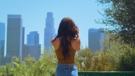Back-view-brunette-tourist-making-video-cityscape-standing-in-green-park.