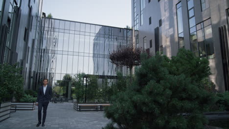 Confident-businessman-walking-office-building.-Landscaped-garden-at-downtown