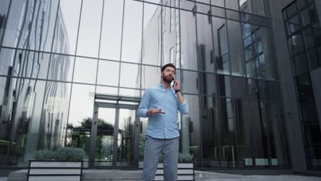 Attractive-man-speaking-phone-at-modern-glass-building.-Manager-working-outside