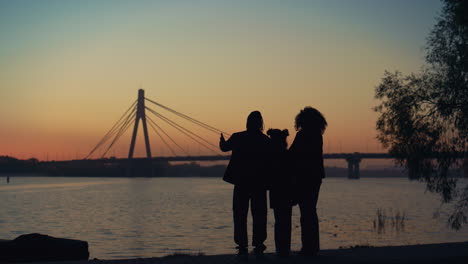 Parents-kid-silhouette-watching-sunset-standing-together-at-river-bridge-view.