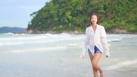 Asian-female-taking-a-walk-along-the-ocean-wearing-blue-shorts-and-a-white-shirt-with-out-of-focus-waves-reaching-the-shore-in-the-background-of-a-tropical-island