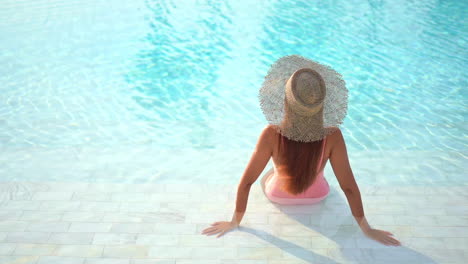 Back-of-Sexy-Female-in-Swimsuit-and-Summer-Hat-Sitting-on-Pool-Edge-on-Hot-Day