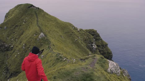 Man-with-red-jacket-walk-on-narrow-path-on-a-cliff-near-the-the-North-Atlantic-ocean-in-stunning-Nordic-landscape-view-on-Kallur-on-the-Faroe-Islands