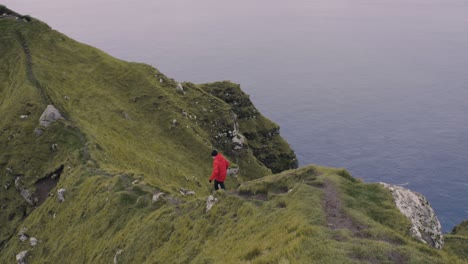Man-in-red-jacket-walks-and-almost-slips-on-narrow-path-on-spectacular-cliff-near-the-North-Atlantic-ocean-in-the-Nordic-landscape-of-Kallur-on-the-Faroe-Islands