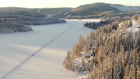 Vista-Aérea-De-Gran-Altitud-De-Un-Lago-Congelado-Y-Esquiadores-De-Fondo-En-Noruega-Con-Puesta-De-Sol-Dorada-Y-Montañas-En-El-Fondo