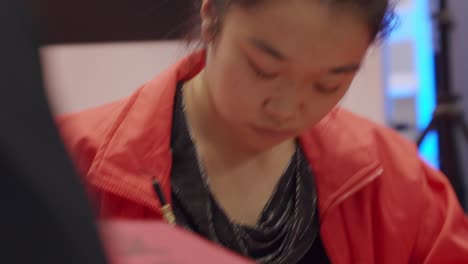 Young-girl,-sitting-at-desk,-writing-Asian-characters,-black-calligraphy,-red-paper