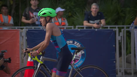 Competidor-Adolescente-Latino-Bajando-De-Su-Bicicleta-Descalzo-Mientras-Ingresa-A-La-Zona-De-Transición-En-Una-Competencia-De-Triatlón-Usando-Un-Casco-Verde