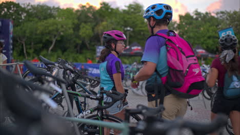 Padre-E-Hija-Colocando-La-Bicicleta-En-El-Estante-En-La-Zona-De-Transición-Preparándose-Para-El-Triatlón-Usando-Cascos-La-Tarde-Antes-De-La-Competencia