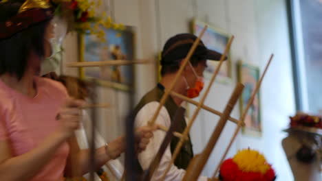 Bamboo-Angklung-being-shaken-by-Asian-woman-wearing-face-mask,-filmed-in-handheld-style-as-medium-close-up-with-panning-motion