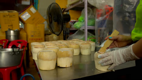 Thailand,-Bangkok---13-August-2022:-Unrecognizable-worker-indoors-industry-backroom-preparing-coconut-bowl-for-coco-jelly-scraping-pulp-using-scraper-tool