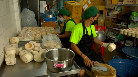 Thailand,-Bangkok---13-August-2022:-Industry-backroom-with-man-cutting-top-of-peeled-green-coconut-and-woman-extracting-coconut-pulp-from-the-shell-using-scraper