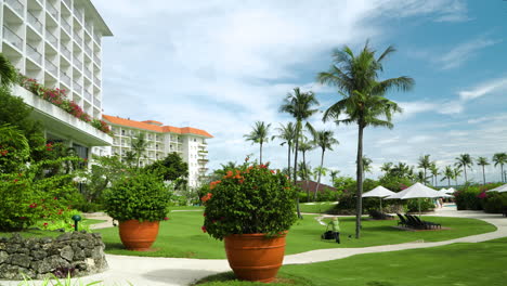 Jardinero-En-El-Hotel-Ocupado-Cortando-El-Césped-En-Un-Día-Soleado-Con-Paisajes-De-Palmeras-Y-Plantas-Florales-En-Macetas