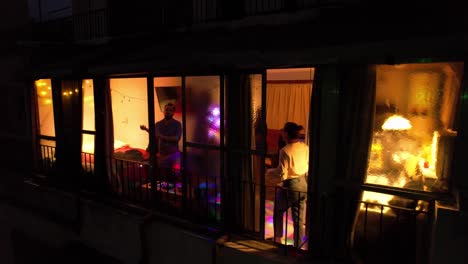 Disparo-De-Un-Dron-Desde-El-Exterior-De-Un-Grupo-De-Personas-Celebrando-Y-Bailando-En-Un-Apartamento-Por-La-Noche,-Luces-Multicolores