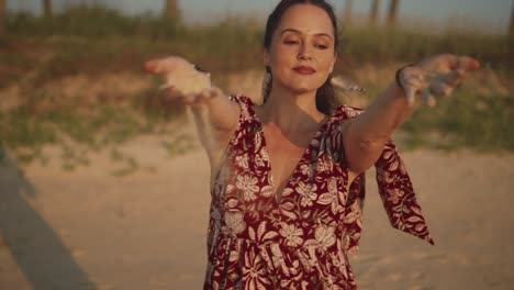 Beautiful-Indian-woman-wearing-a-red-summer-dress,-letting-sand-blow-in-the-wind-from-her-hands,-on-a-tropical-beach