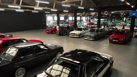 Club-fans-admiring-classic-BMW-e30-vehicles-parked-in-warehouse-meeting-exhibition,-aerial-reveal-view