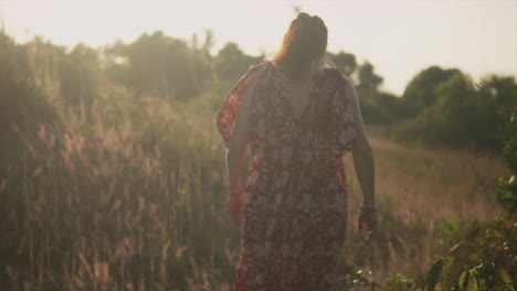 Beautiful-brunette-Indian-woman-walking-away-through-high-grass-and-trees,-wearing-a-red-summer-dress,-on-a-sunny-evening