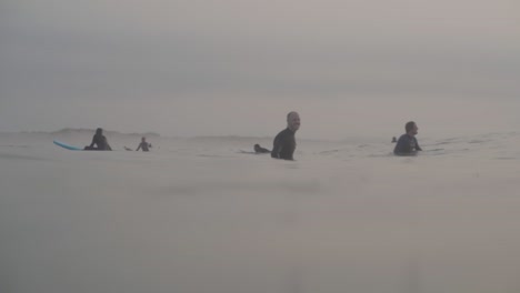 Grupo-De-Surfistas-Esperando-El-Oleaje,-Surfista-En-Primer-Plano-Pasando-Remando,-Peniche-Matutino-Nublado