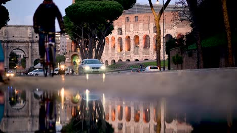 Coches,-Bicicletas-Y-Scooters-Pasando-Por-El-Reflejo-En-Un-Charco-Fuera-Del-Coliseo,-Roma,-Italia.