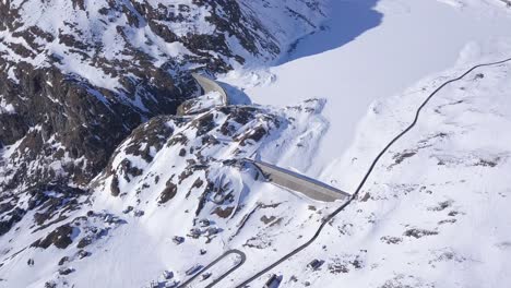 Die-Langsame-Neigung-Der-Drohne-Zeigt-Die-Schneebedeckten-Italienischen-Berge-Im-Winter,-Den-Schneebedeckten-See-Und-Kurvenreiche-Straßen-Rund-Um-Den-Damm