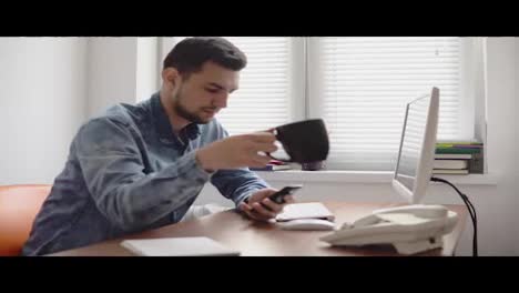 Young-office-worker-using-his-phone-at-the-office-sitting-at-the-table-with-computer,-phone-and-drinking-coffee-or-tea-from-his