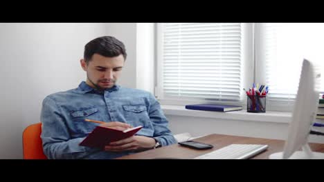 Young-businessman-sitting-by-the-computer-in-stylish-modern-office-and-taking-notes-using-his-pencil-and-notebook.-Computer