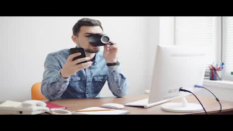 Young-office-worker-using-his-phone-at-the-office-sitting-at-the-table-with-computer,-phone-and-drinking-coffee-or-tea-from-his