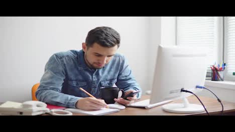 Joven-Empresario-Sentado-Frente-A-La-Computadora-En-Una-Elegante-Oficina-Moderna-Y-Tomando-Notas-Con-Lápiz-Y-Teléfono.-Computadora,-Teléfono