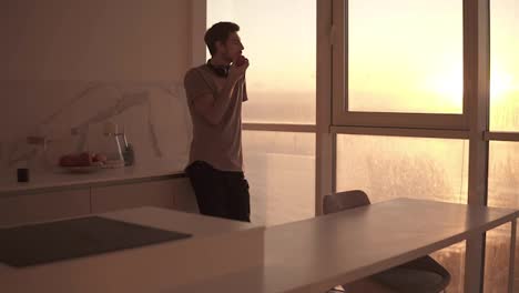 Tall,-young-single-man-with-headphones-on-neck-relaxing-by-the-window-at-studio-kitchen,-watching-sunset-and-taking-apple-in-his