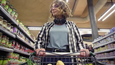 Una-Mujer-Alegre-Con-Camisa-A-Cuadros-Y-Auriculares-En-El-Cuello-Camina-En-Una-Tienda-De-Comestibles-Dirigiendo-Un-Carrito-De-Compras-Con-Comida-En-Su-Interior