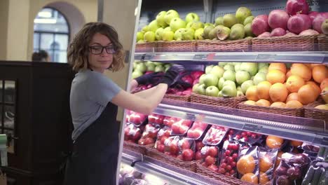 Close-up-of-pretty-caucasian-worker-in-black-apron-and-gloves-stocking-the-fruits-in-supermarket.-Young-employee-in-glasses-at