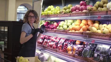 Cerca-De-Un-Trabajador-Bastante-Caucásico-Con-Delantal-Negro-Y-Guantes-Almacenando-Las-Frutas-En-El-Supermercado.-Joven-Empleado-Con-Gafas-En