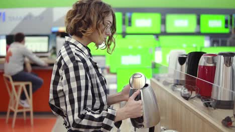 Vista-Lateral-De-Una-Joven-Alegre-Y-Atractiva-Mujer-Con-Pantalones-A-Cuadros-Eligiendo-Un-Hervidor-Electrónico-En-Una-Tienda-De-Electrodomésticos,-Parada-En-Fila,-Sosteniendo-Y-Examinando-Un-Hervidor-De-Plata