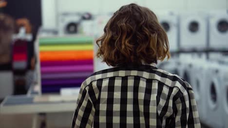 Tracking-rare-view-footage-of-a-girl-in-a-plaid-shirt-and-short-hair-walking-by-supermarket-in-household-appliences-looking-for-something.-Blurred-background