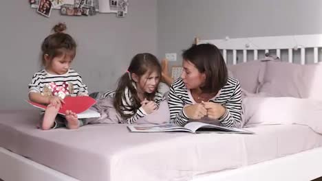 Young-loving-mother-and-daughters-are-reading-a-book-and-discussing-while-lying-on-bed-in-bedroom-at-home