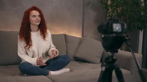 Curly-redhead-young-girl-vlogger-is-talking-in-front-of-camera-recording-video-for-online-blog-in-internet-speaking,-smiling