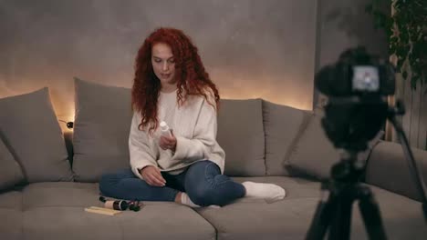 Curly-redhead-young-girl-vlogger-is-talking-in-front-of-camera-recording-video-for-online-blog-in-internet,-demosrating