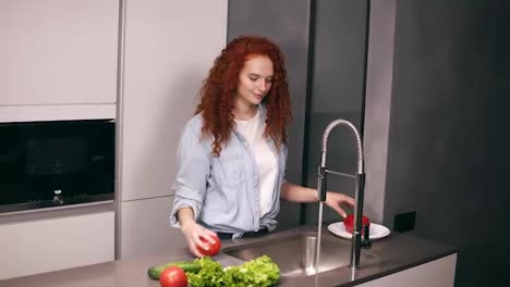 Charming-redhead-woman-washing-red-pepper,-cucumbers-and-tomato-in-kitchen-and-putting-them-on-plate.-Homemade-food-concept