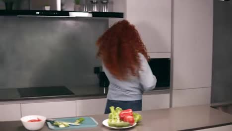 Happy-active-redhead-young-woman-holding-spoon-as-a-microphone-singing-songs-and-dancing-while-cooking-in-kitchen,-carefree