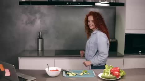 Happy-active-redhead-young-woman-holding-spoon-as-a-microphone-singing-songs-and-dancing-while-cooking-in-kitchen,-carefree