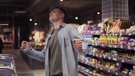 At-the-supermarket:-Happy-stylish-man-with-dances-freely-in-big-shopping-mall-in-front-the-shelves-with-goods.-Positive-dances