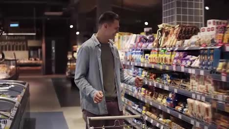 At-the-supermarket:-Happy-stylish-man-with-shopping-trolley-in-big-shopping-mall-walking-by-the-shelves-with-goods-and-suddenly