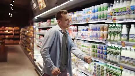 Smiling,-stylish-man-with-shopping-trolley-in-big-shopping-mall-walking-by-the-shelves-with-milk-products-and-suddenly-starts