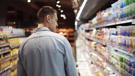 and-attractive-tall-guy-in-supermarket.-Rare-view-caucasian-young-man-in-blue-shirt-looking-for-a-sour-cream-and-take-it-from-the