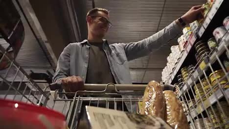 A-positive-young-guy-in-glasses-putting-goods-in-shopping-trolley.-Passing-through-the-canned-goods-aisle-of-a-big-supermarket