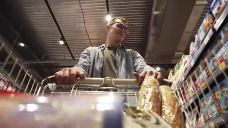 A-cheerful-young-man-in-glasses-putting-goods-in-shopping-trolley.-Passing-through-the-dry-goods-aisle-of-a-big-supermarket.-Low