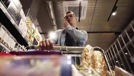 At-the-Supermarket:-Handsome-man-with-shopping-cart-uses-smartphone---talking.-Looks-at-dairy-products-and-walks-through-dry