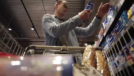 Male-shopping.-A-man-in-glasses-putting-goods-in-shopping-trolley.-Passing-through-the-dry-goods-aisle-of-a-big-supermarket.-Low