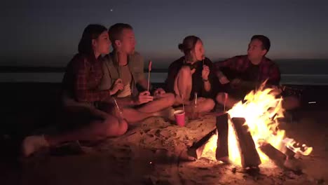 Un-Grupo-De-Cuatro-Amigos-Se-Reúnen-En-La-Playa-Cerca-De-La-Fogata,-Comiendo-Salchichas,-Bebiendo-Y-Tocando-La-Guitarra-En-Un-Mar-Oscuro.