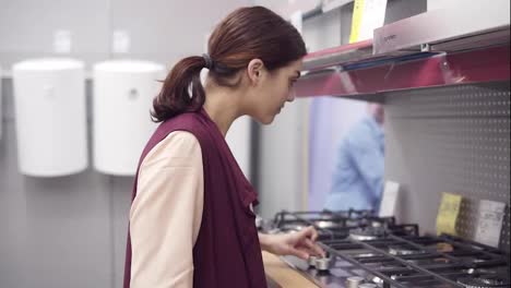 Young-brunette-girl-presses-burners-buttons-on-cooktop-of-kitchen-stove-and-takes-the-price-tag-from-display-in-hardware-store.-Choosing-household-appliance-equipment.