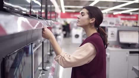 Una-Clienta-Joven-Que-Elige-Un-Nuevo-Televisor-Compara-Los-Precios-De-Diferentes-Pantallas-De-Televisión-En-El-Departamento-De-Electrónica-De-Una-Tienda-De-Electrodomésticos.